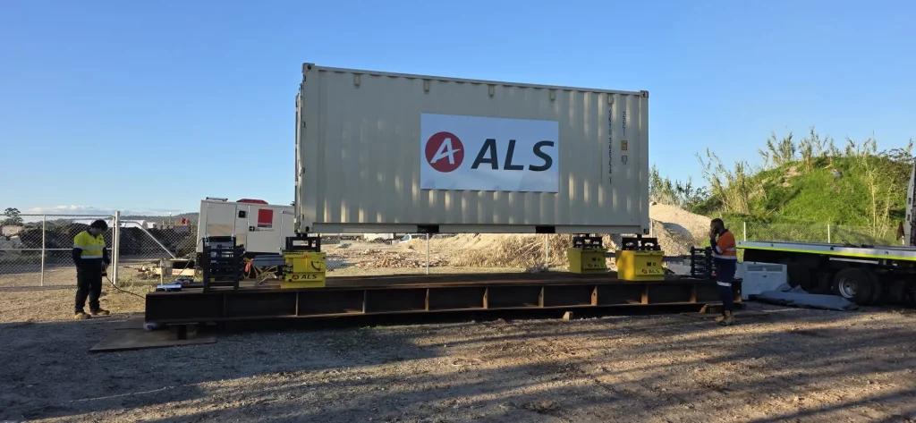 ALS International branded shipping container on a steel transport frame with riggers in hi-vis