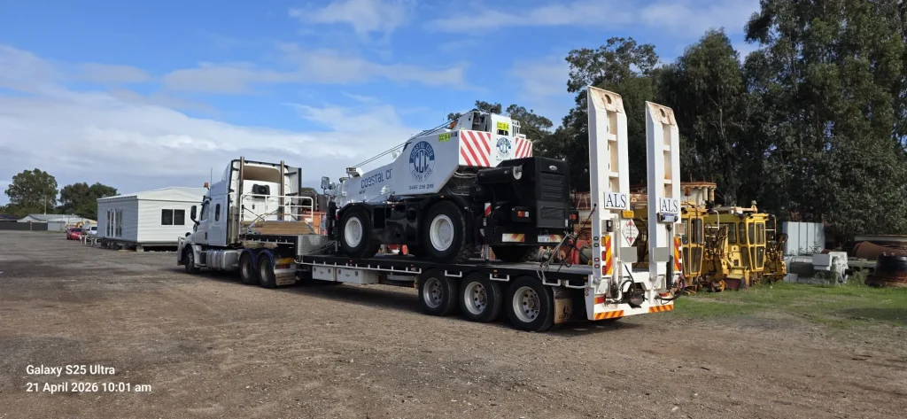 ALS International prime mover hauling a mobile crane on a float at a rural depot