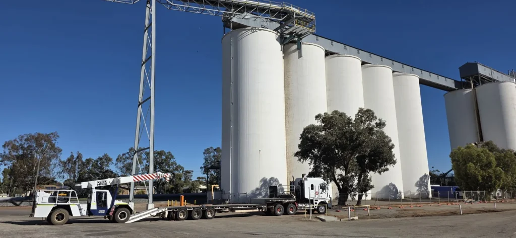 Mobile crane loaded on an ALS International low-loader float beside grain storage silos